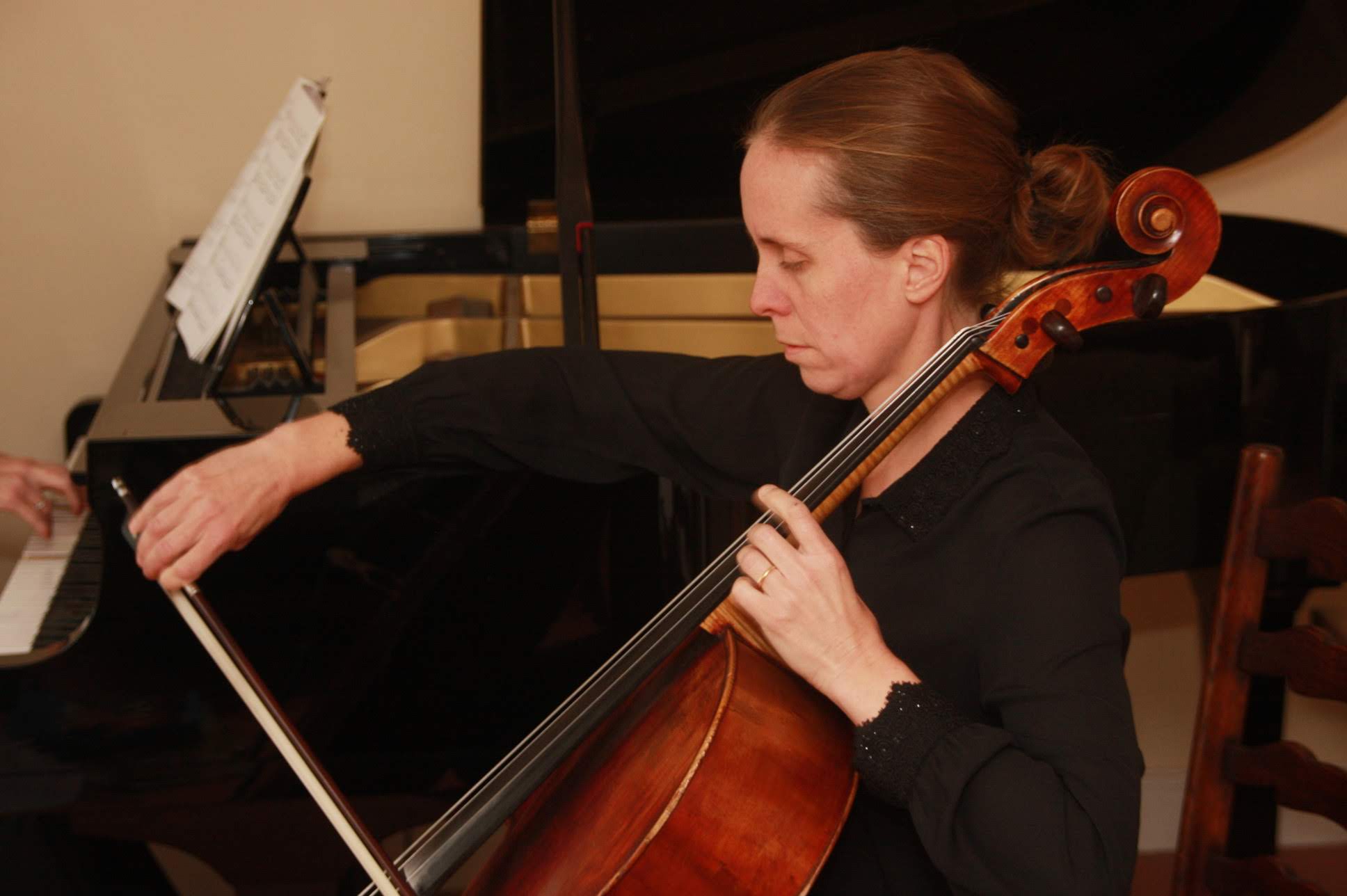 A woman with brown hair and a bun, playing a cello in front of a piano.