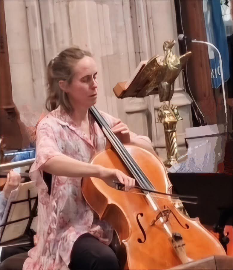 A woman plays the cello, seated and attired in black pants and an unbuttoned, long-sleeved pink tie-dye blouse, fingers po...